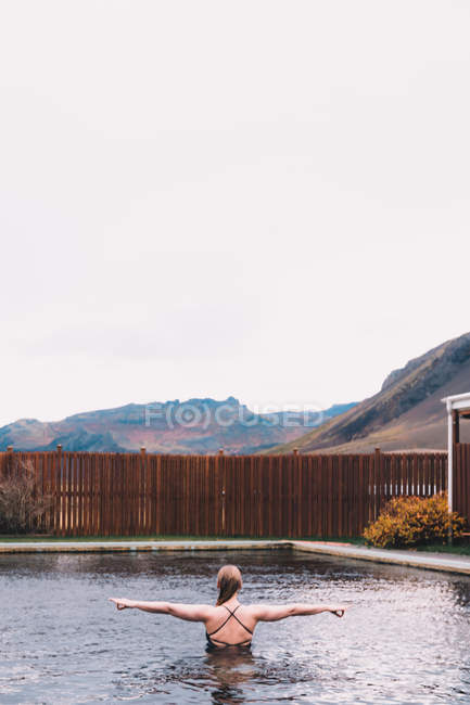 Vista posterior de la joven mujer descansando en el agua de la piscina contra la valla de madera en la naturaleza con la montaña en el fondo - foto de stock