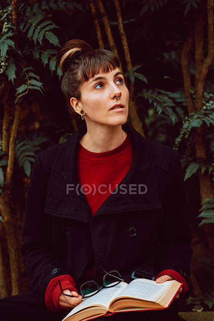 Young dreamy woman with piercing holding book on bench in city garden — Stock Photo