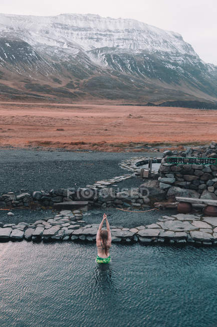 Обратный вид женщины с поднятыми руками плавающей в воде возле скал на берегу и горы в снегу между дикими землями — стоковое фото