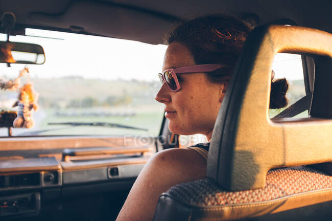 Vista trasera de una mujer bastante joven en gafas de sol mirando hacia otro lado mientras está sentada en el asiento del pasajero de un coche moderno durante un viaje por carretera en Cantabria, España - foto de stock