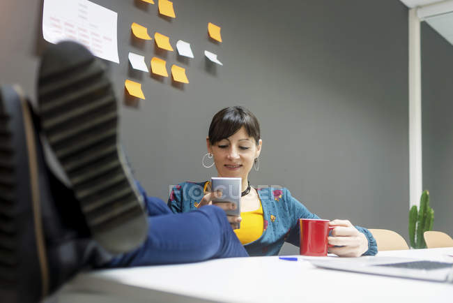 Manager donna con tazza di bevanda calda utilizzando lo smartphone mantenendo le gambe sulla scrivania in ufficio — Foto stock
