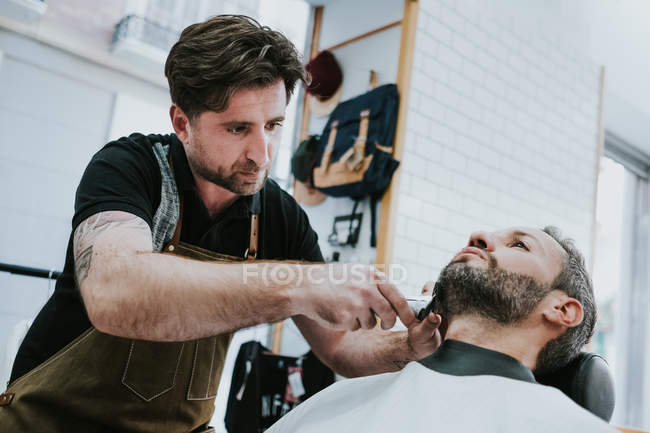 Barbiere con pettine e trimmer taglio barba di maschio seduto in barbiere su sfondo sfocato — Foto stock