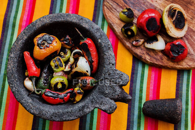 Köstliches gebratenes Gemüse im Mörser und auf Holzbrettern auf bunt gestreifter Tischdecke — Stockfoto