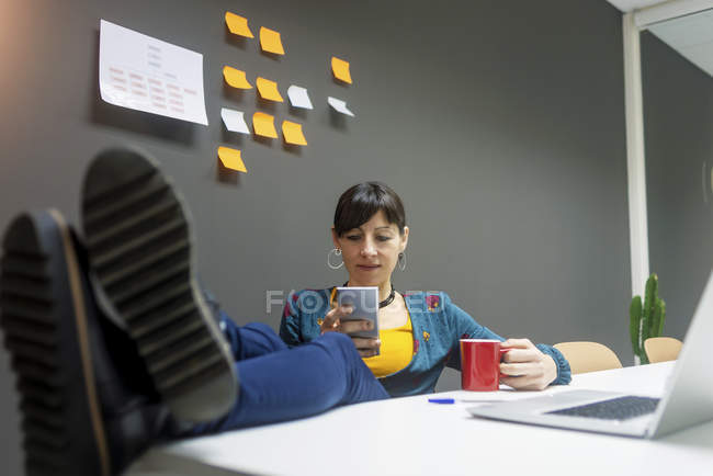Female manager with mug of hot beverage using smartphone while keeping legs on desk in office — Stock Photo
