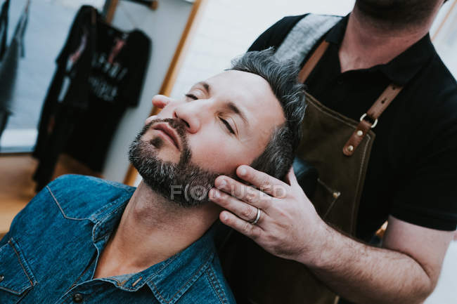 Barbeiro fazendo massagem facial para bonito macho elegante com olhos fechados sentado na cadeira — Fotografia de Stock