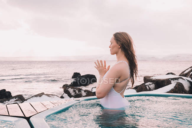 Молода жінка з закритими очима роздумує у воді басейну біля скель і хмарного неба — стокове фото