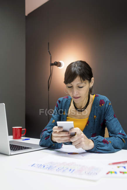 Mulher com caneca de bebida quente usando smartphone perto do laptop enquanto trabalhava no escritório moderno — Fotografia de Stock