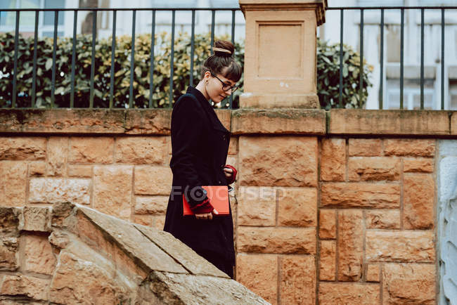 Молодая элегантная женщина в очках держит книгу и спускается по лестнице перед зданием в городе — стоковое фото