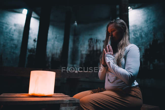 Vista lateral da jovem com as mãos de oração sentadas perto de luzes no prédio escuro — Fotografia de Stock