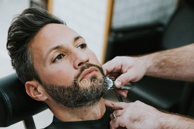 Closeup of barber with comb and trimmer cutting beard of male sitting in barbershop on blurred background — Stock Photo
