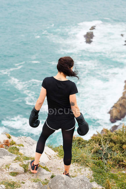 Rückansicht einer Frau in Boxhandschuhen, die auf einer steinigen Klippe steht und beim Outdoor-Training aufs Meer blickt — Stockfoto