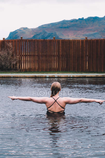 Rückansicht einer jungen Frau, die sich im Wasser des Schwimmbeckens ausruht, gegen einen Holzzaun in der Natur mit einem Berg im Hintergrund — Stockfoto