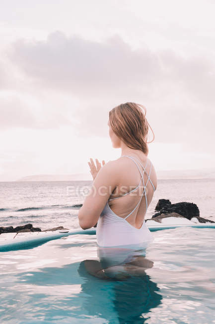 Mulher jovem meditando na água da piscina perto de rochas e céu nublado — Fotografia de Stock