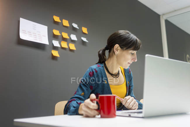 Geschäftsfrau mit einem Becher Heißgetränk surft im Laptop, während sie im modernen Büro arbeitet — Stockfoto