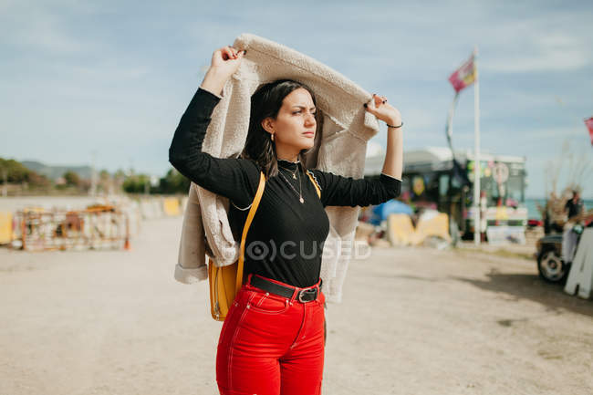 Junge brünette Frau im trendigen Outfit bedeckt Kopf mit Jacke und schaut weg, während sie auf verschwommenem Hintergrund der Küste steht — Stockfoto