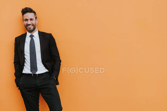 Взрослый красивый элегантный бизнесмен в официальном костюме смотрит в камеру возле оранжевой стены — стоковое фото