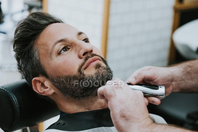 Close-up de barbeiro com pente e cortador de barba de corte de macho sentado na barbearia em fundo borrado — Fotografia de Stock