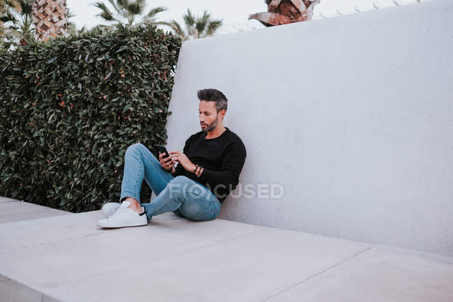 Gutaussehender eleganter glücklicher Mann mittleren Alters in Freizeitkleidung, der ein Mobiltelefon benutzt und in der Nähe einer grauen Wand sitzt — Stockfoto