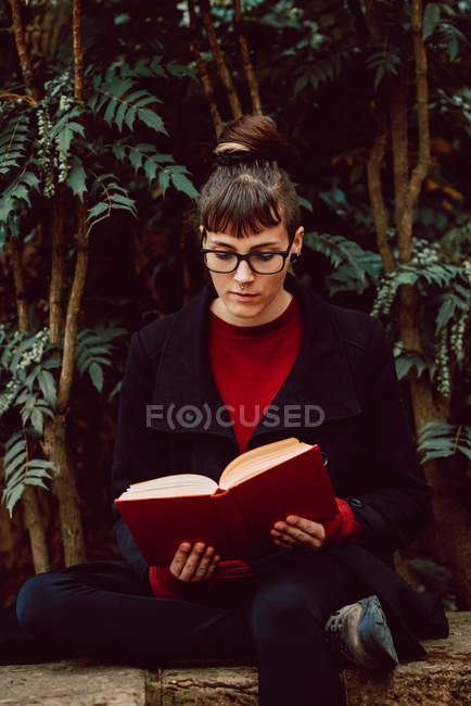 Young elegant woman in eyeglasses reading book and sitting on bench in city garden — Stock Photo