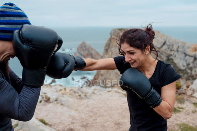 Man and woman in boxing gloves punching each other while standing on cliff against sea and sky — Stock Photo