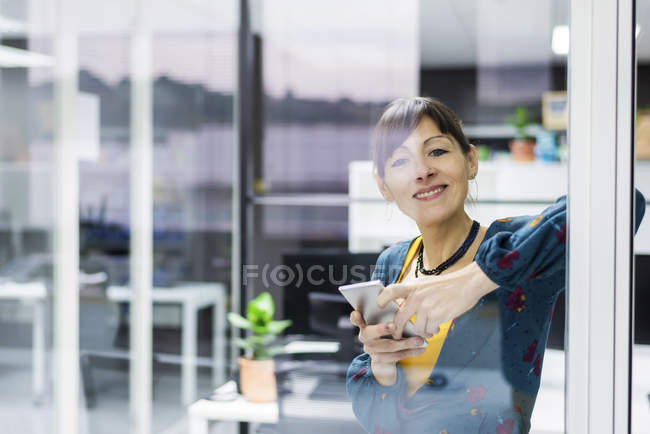 Gestora sonriente sosteniendo teléfono inteligente mientras está de pie cerca de la pared de vidrio en la oficina moderna - foto de stock