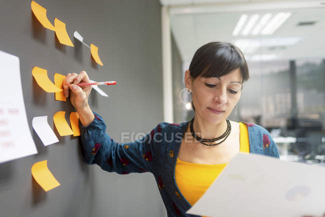 Бізнес-леді тримає документ і пише на липких нотах, стоячи біля сірої стіни в офісі — стокове фото