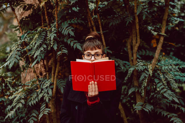 Молода елегантна жінка в окулярах дивиться на камеру, прикриваючи обличчя книгою в міському саду — стокове фото