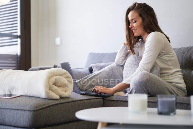 Весела молода жінка, що використовує ноутбук і відпочиває на дивані вдома — стокове фото