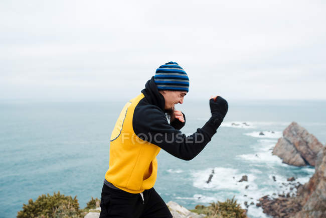 Uomo barbuto adulto in abbigliamento sportivo che pratica pugni durante l'allenamento di kickboxing sulla scogliera rocciosa vicino al mare — Foto stock