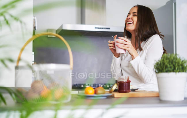 Young laughing woman holding mug and having breakfast in modern kitchen at home — Stock Photo