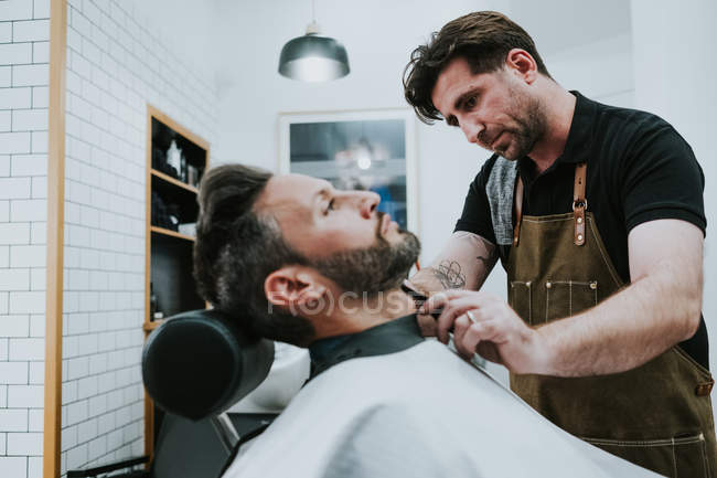 Friseur mit Kamm und Trimmer schneiden Bart des Mannes sitzt in Friseursalon auf verschwommenem Hintergrund — Stockfoto
