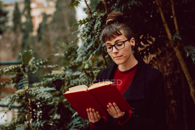 Молода приваблива елегантна жінка в окулярах читає книгу в міському саду — стокове фото