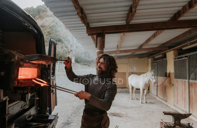 Bearded adult farrier taking hot horseshoe from portable furnace in back of car while working near stable on ranch — Stock Photo