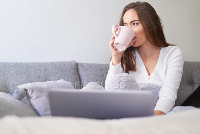 Молода жінка п'є з чашки на дивані з ноутбуком вдома — стокове фото