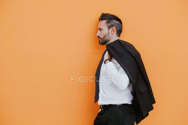 Vista lateral de adulto guapo elegante hombre de negocios alegre en traje formal con la mano en el bolsillo mirando hacia otro lado cerca de la pared naranja en la calle de la ciudad - foto de stock