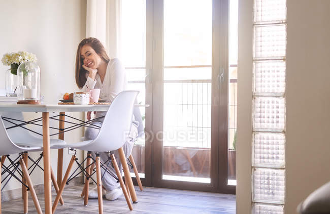Jovem mulher feliz atraente tomando café da manhã à mesa perto da janela em casa — Fotografia de Stock