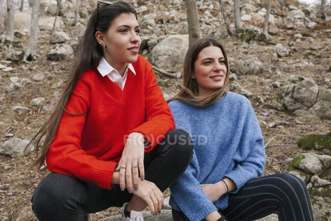Young attractive happy friends in bright knitted sweaters looking away and sitting on stones in forest — Stock Photo