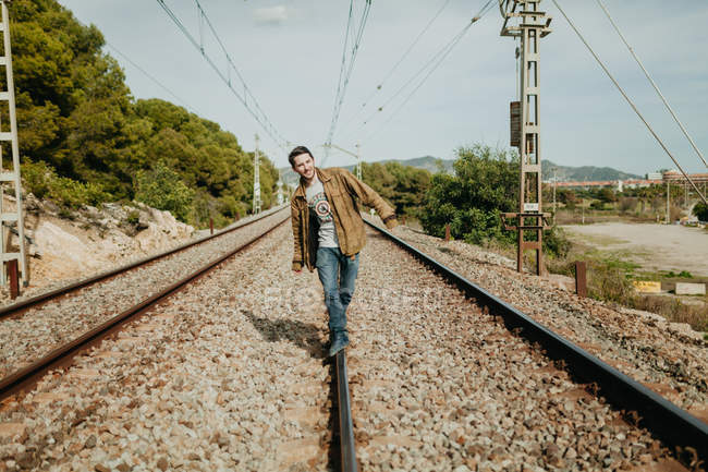 Молодой веселый мужчина в повседневной одежде смотрит в камеру во время прогулки по рельсам поезда — стоковое фото