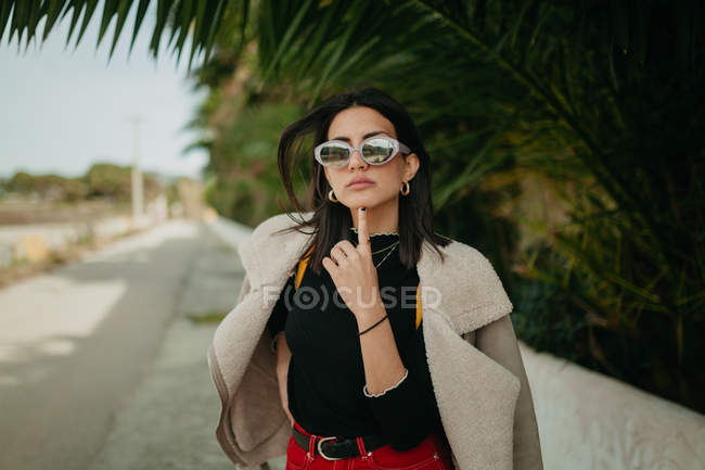 Стильна молода жінка в сонцезахисних окулярах стоїть біля листя тропічної долоні на вулиці — стокове фото