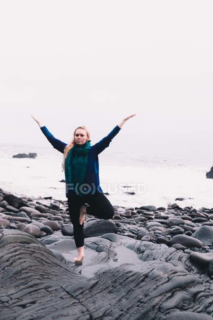 Young woman with upped hands meditating and standing on rocks near sea coast — Stock Photo