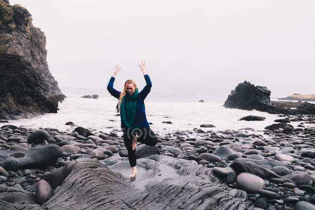 Jeune femme avec les mains levées méditant et debout sur des rochers près de la côte de la mer — Photo de stock