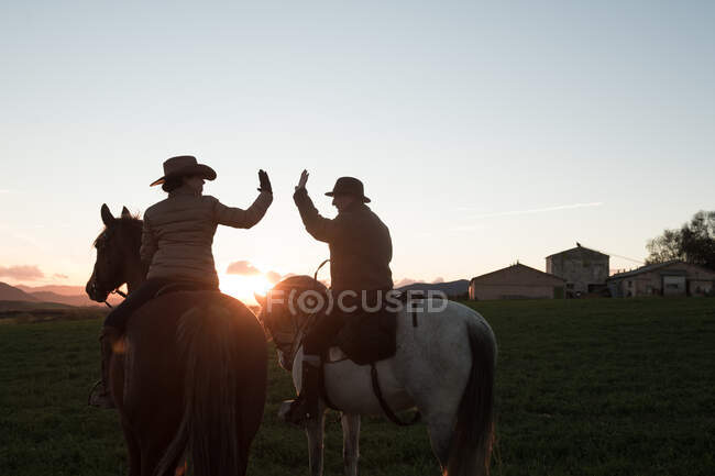 Vista posteriore di uomo e donna che cavalcano cavalli e si danno cinque a vicenda contro il cielo al tramonto nel ranch — Foto stock