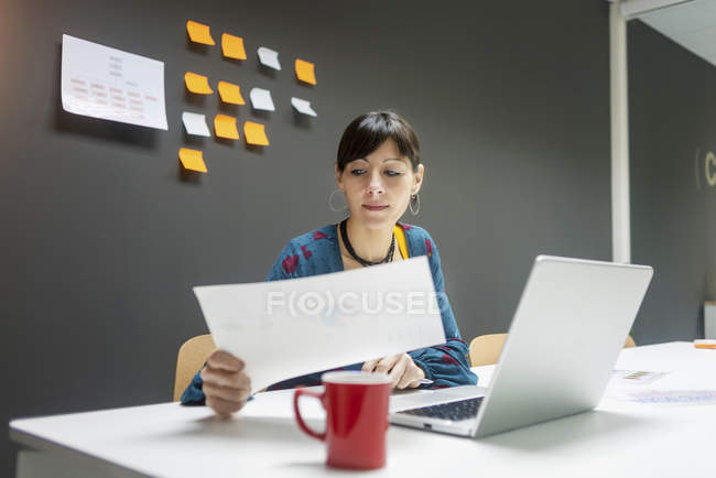 Donna d'affari che lavora con documenti mentre è seduta alla scrivania con computer portatile in ufficio moderno — Foto stock