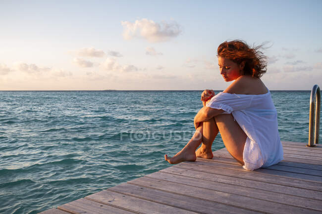 Вид сбоку на красивую молодую женщину, смотрящую вниз, сидя на деревянном пирсе возле волнистой морской воды на фоне облачного вечернего неба на Мальдивах — стоковое фото