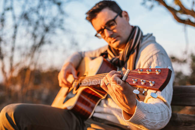 Casual uomo in occhiali suonare la chitarra su panchina in campagna — Foto stock