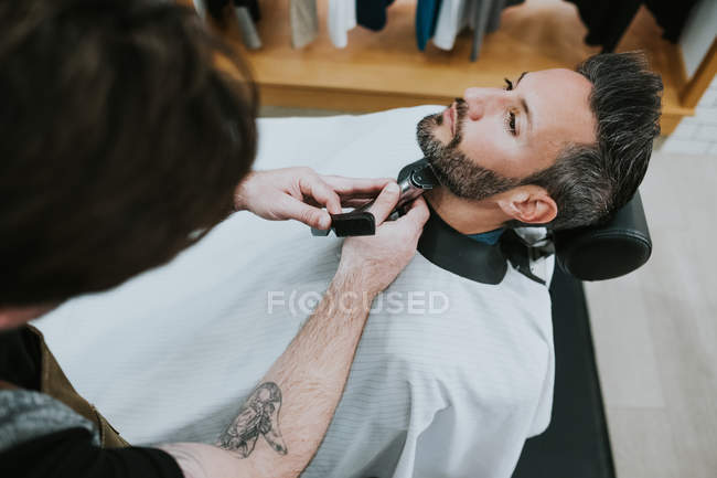 Peluquero con peine y recortadora barba de corte de macho sentado en la barbería sobre fondo borroso - foto de stock