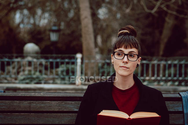 Jovem mulher elegante em óculos com livro sentado no banco no parque da cidade — Fotografia de Stock