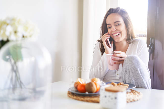 Giovane donna felice che parla al cellulare mentre fa colazione a tavola vicino alla finestra di casa — Foto stock