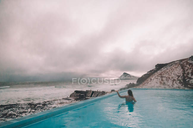 Вид ззаду молода жінка підняла руку, відпочиваючи у воді басейну біля скель на узбережжі і штормового моря — стокове фото