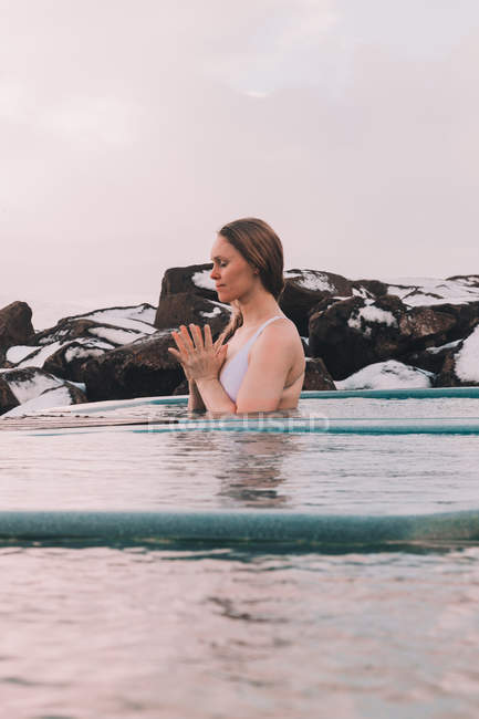 Jovem com olhos fechados meditando na água da piscina perto de rochas e céu nublado — Fotografia de Stock